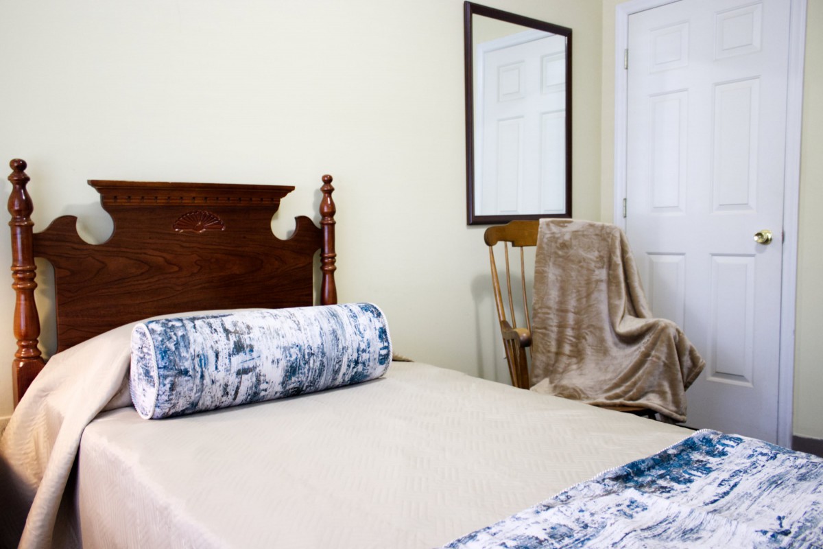 A cozy bedroom with a neatly made bed, a decorative pillow, and a wooden chair draped with a blanket beside a mirror on the wall.