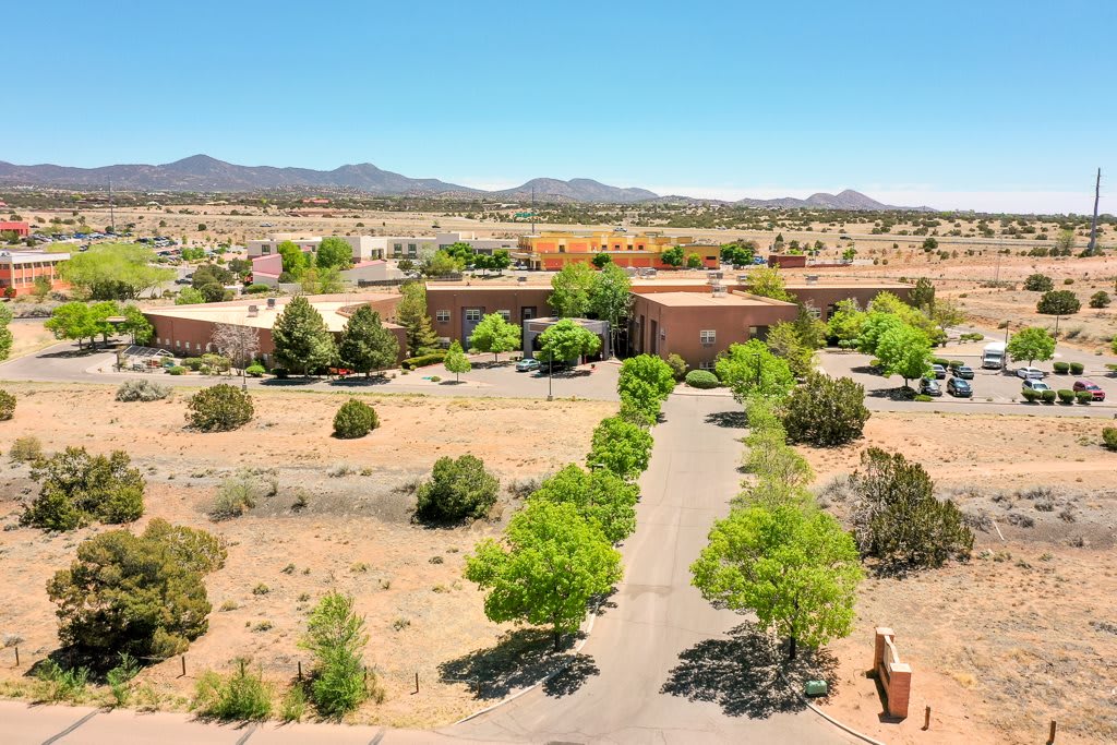 Aerial view of a desert landscape with a building surrounded by trees and a road leading up to it, set against a backdrop of mountains and clear blue sky.