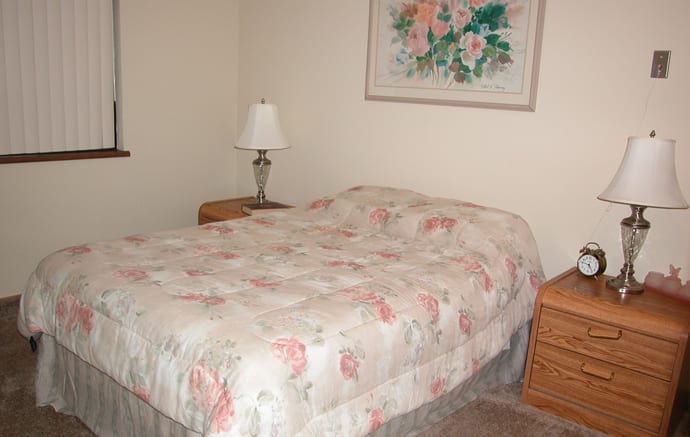 A cozy bedroom featuring a bed with a floral-patterned blanket, two wooden nightstands, and a flower painting above the bed.