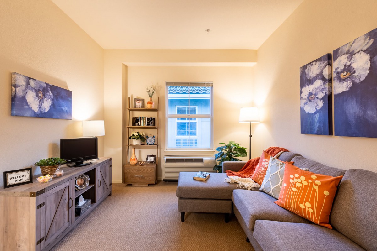 A cozy living room featuring a sofa with colorful cushions, a TV stand, bookshelf, and warm lighting.