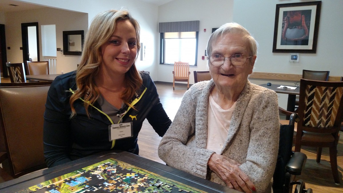 A caregiver sits next to a smiling elderly woman in an indoor setting with a puzzle on the table.