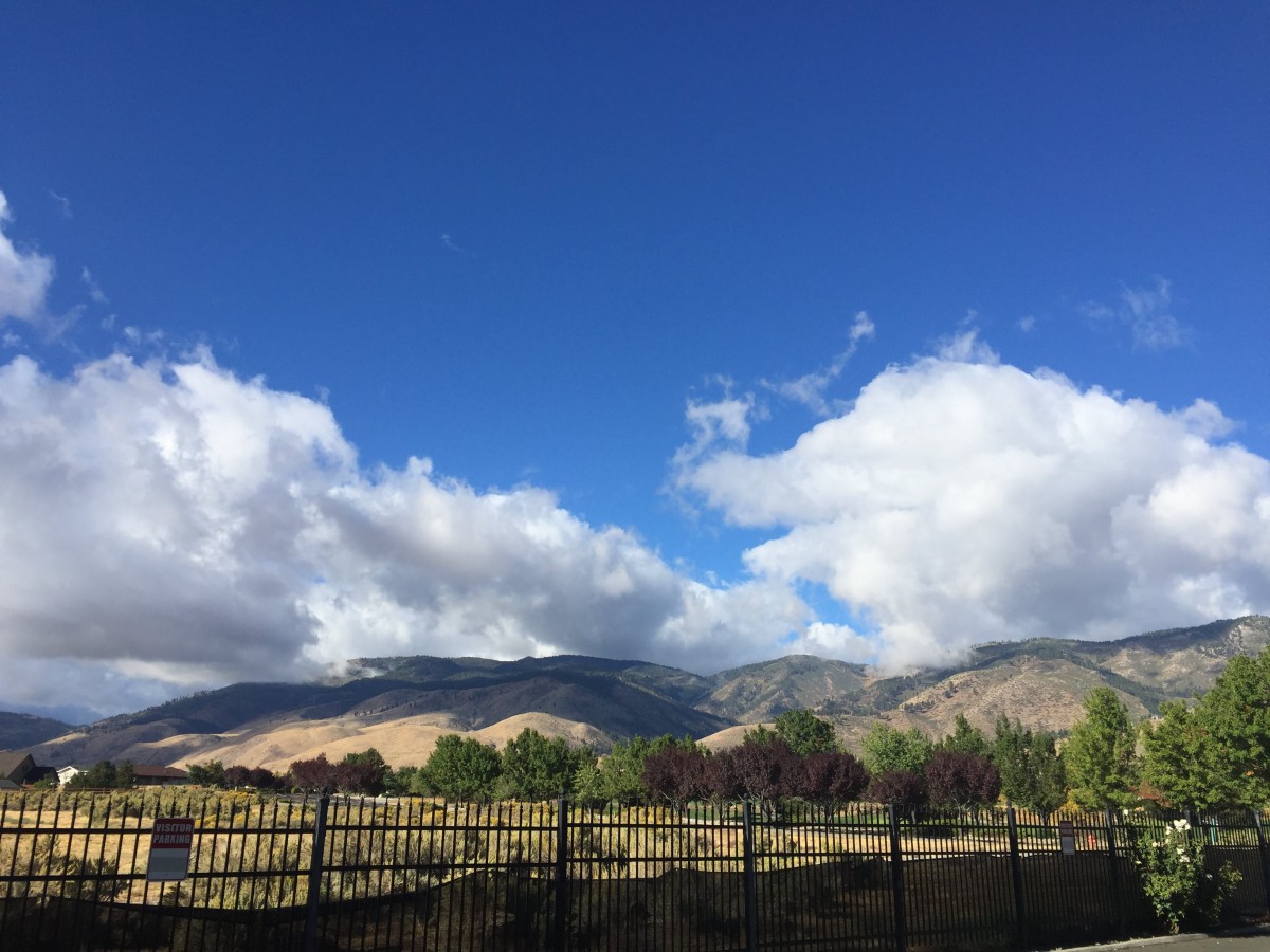 A scenic view of mountains under a bright blue sky with fluffy clouds and a wooden fence in the foreground.