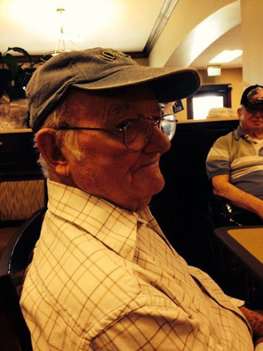 An elderly man wearing a cap and glasses, seated indoors with a relaxed expression.