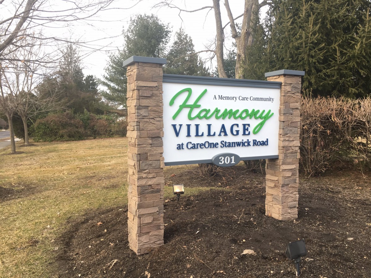 Entrance sign for Harmony Village at CareOne Stanwick Road surrounded by outdoor greenery.