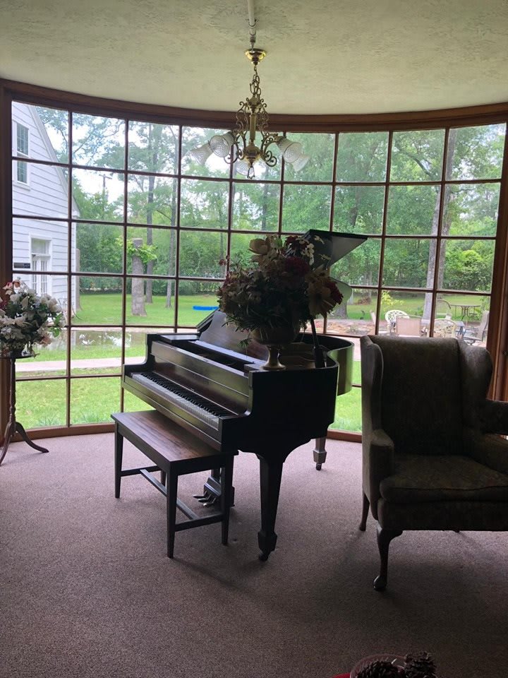 A grand piano sits in a bright living room with large windows overlooking a garden.