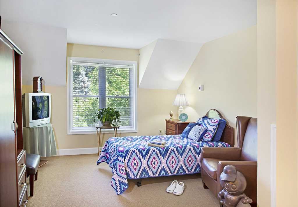 A cozy bedroom featuring a single bed, armchair, and television with natural light pouring in through a large window.