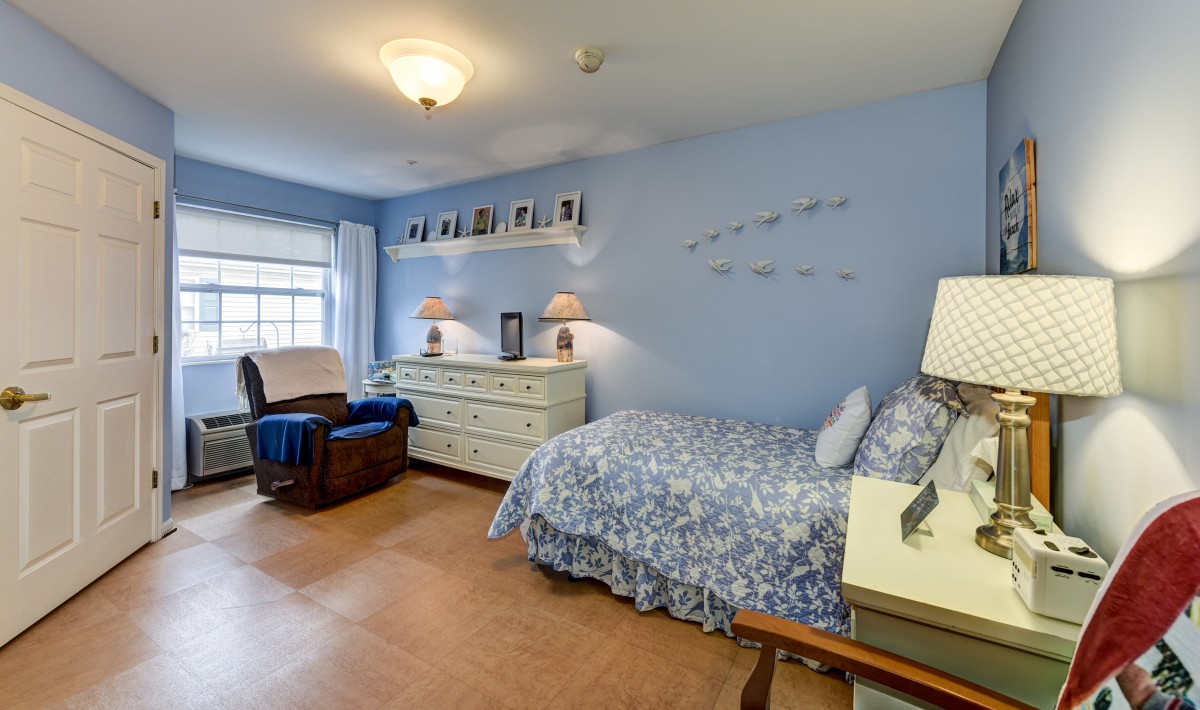 A cozy bedroom with blue walls features a single bed, dresser, and a comfortable chair by the window.