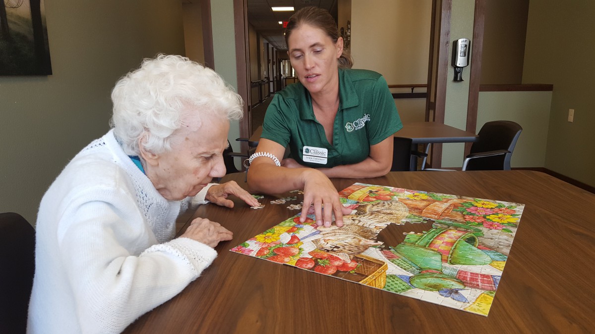 A caregiver assists an elderly woman in completing a colorful puzzle at a nursing home.