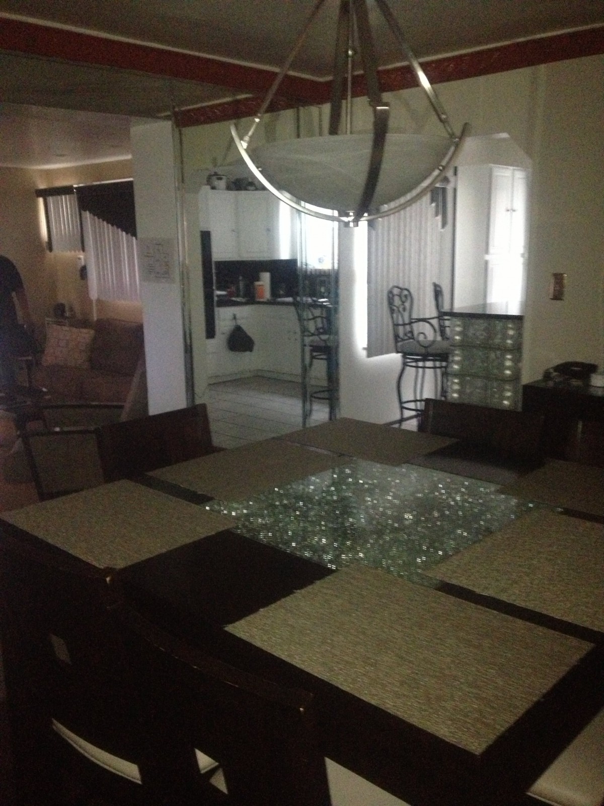 A dining room with a glass table and surrounding chairs, with a view into the kitchen area.