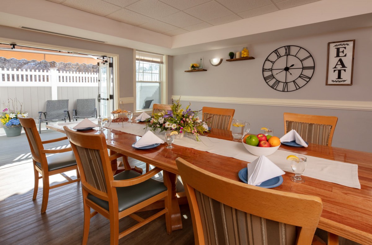 A cozy dining room with a wooden table set for a meal, featuring a decorative clock and shelves on the wall.