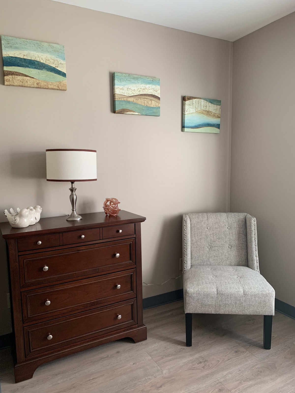 A cozy corner with a wooden dresser, a small chair, and decorative paintings on the wall.