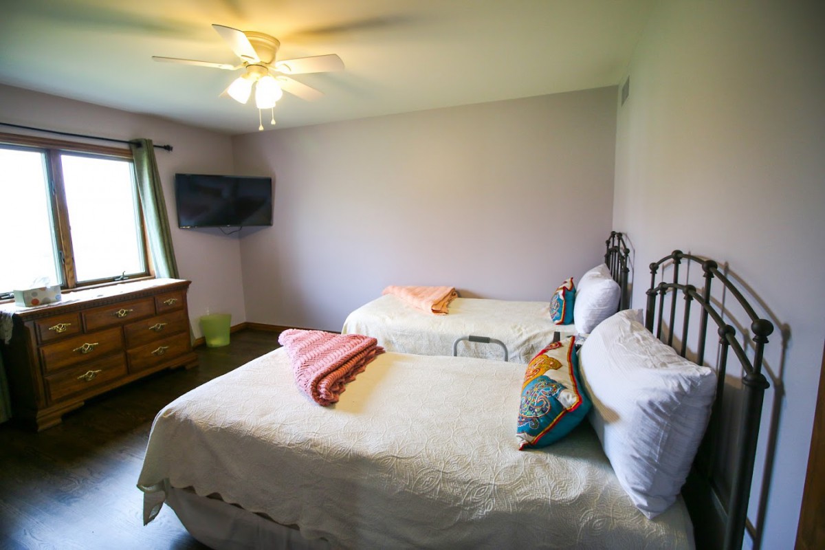 A bright bedroom with two twin beds, a ceiling fan, and a wall-mounted television.