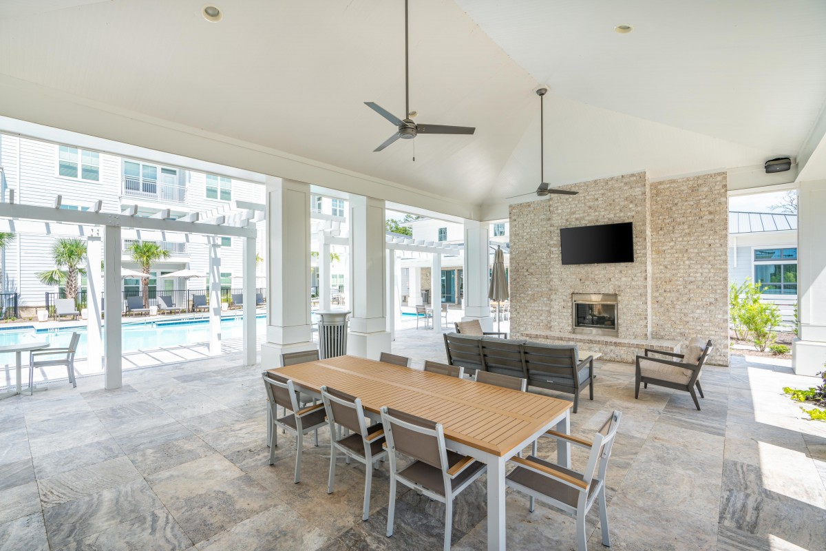 A modern indoor dining area with a wooden table, chairs, and ceiling fans, featuring a fireplace and television, with large windows overlooking a pool.