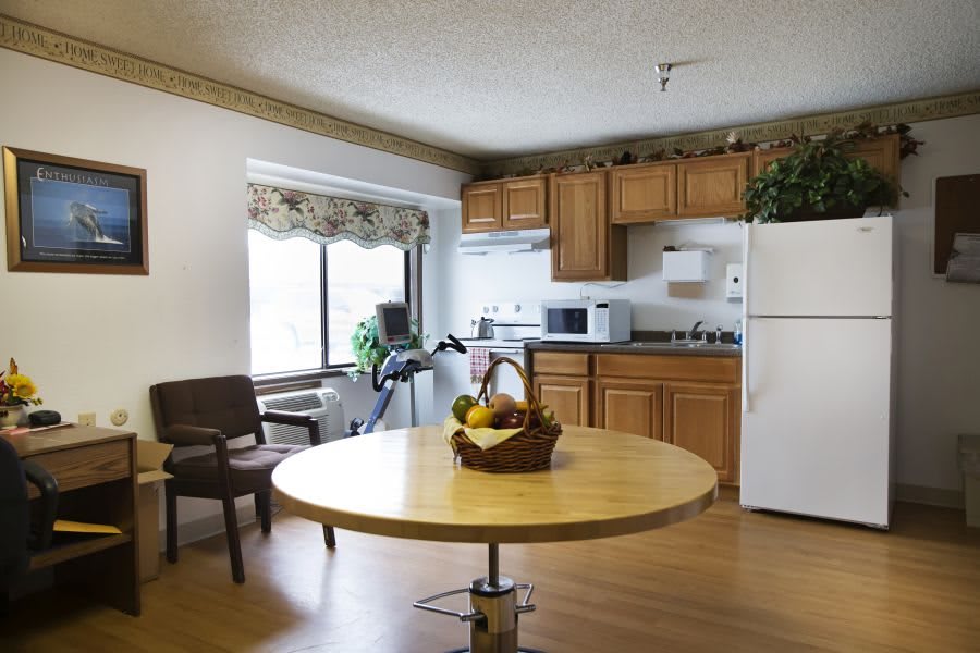 A cozy kitchen and dining area with a round table, chairs, and a fruit basket. The space includes wooden cabinets, a refrigerator, microwave, and is well-lit by a large window.