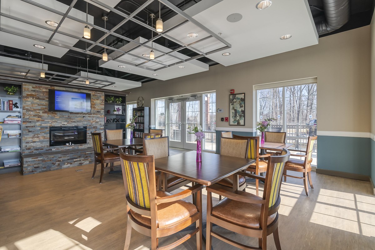A modern dining area with tables, chairs, a television, and a fireplace, featuring large windows that let in natural light.