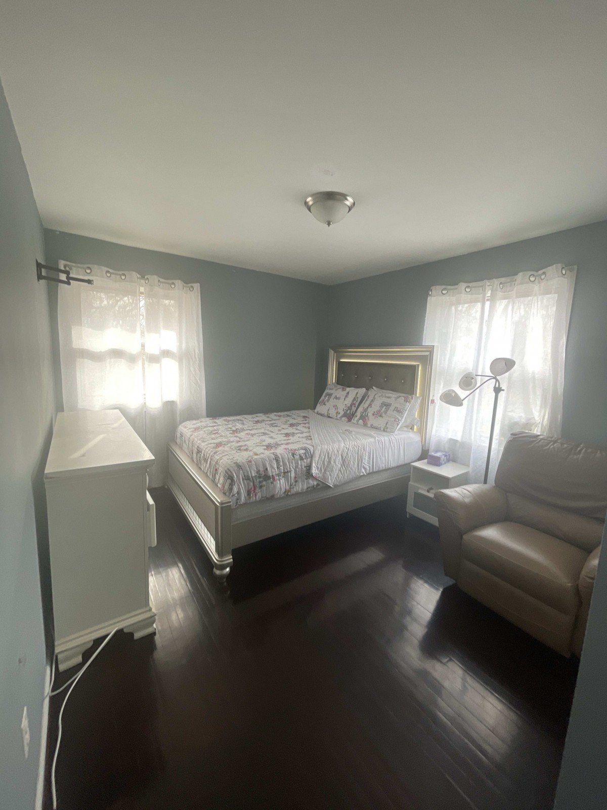 A cozy bedroom with a bed, armchair, and dresser, featuring two windows with sheer curtains.