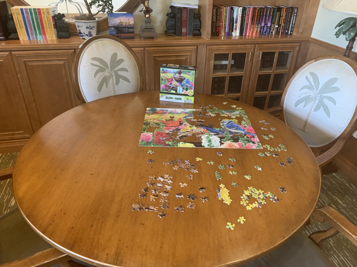 An incomplete jigsaw puzzle on a round wooden table with a bookshelf in the background.