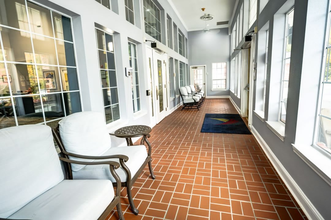 A bright hallway with large windows, featuring cushioned chairs and a tiled floor.