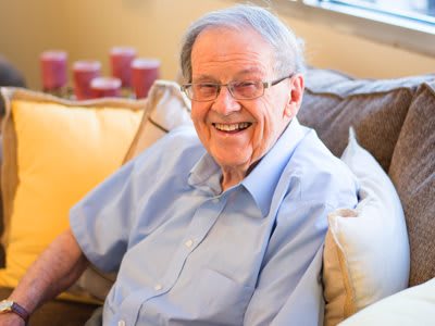An elderly man sitting comfortably on a couch, smiling warmly.