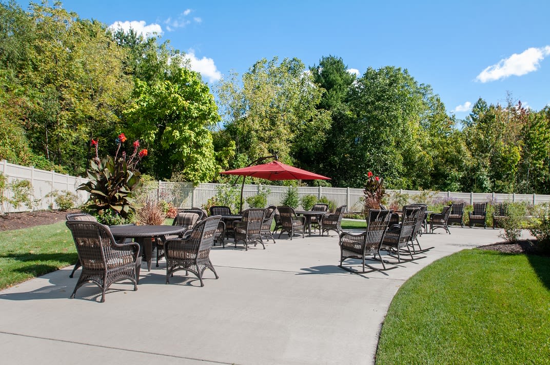 A sunny outdoor patio with tables, chairs, and a red umbrella surrounded by lush greenery.