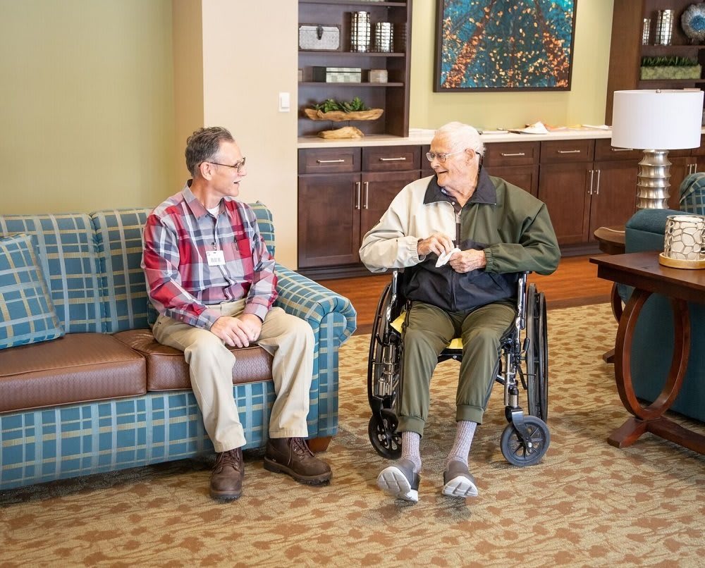 Two elderly men chatting in a comfortable living room setting, one seated on a couch and the other in a wheelchair.