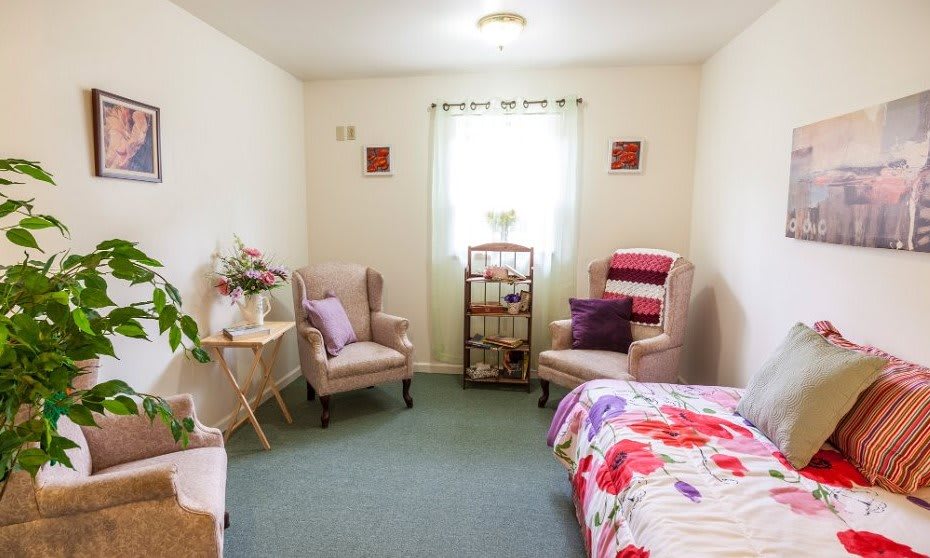 A cozy bedroom with two armchairs, a bed with floral bedding, and bright natural light.
