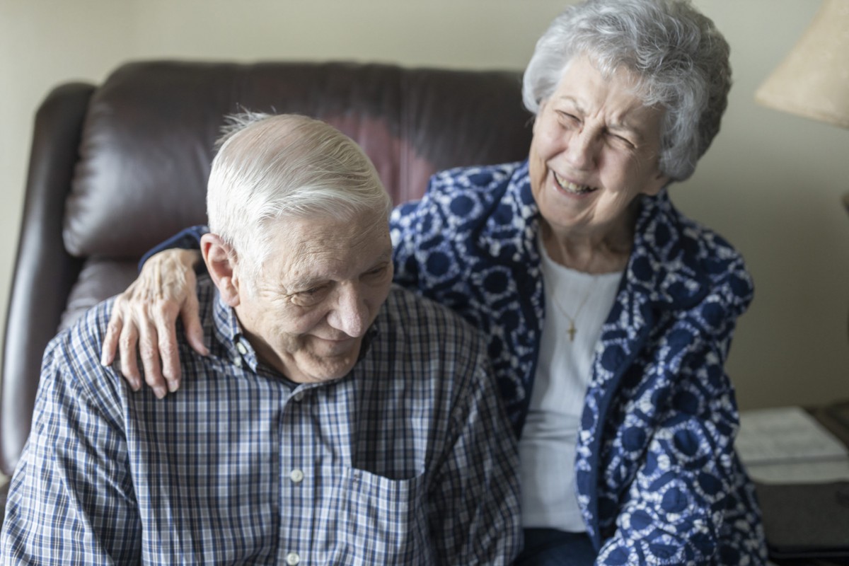 An elderly couple sitting together on a couch, smiling warmly.
