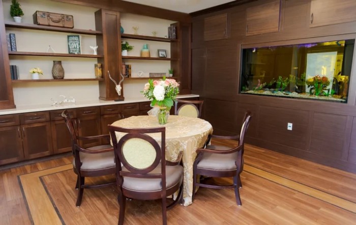 A cozy dining room with a round table and floral centerpiece, surrounded by elegant chairs. The room includes wooden flooring, built-in shelves with decorative items, and an aquarium embedded in the wall.