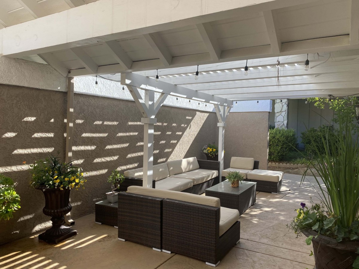 A modern outdoor patio with a pergola, featuring comfortable seating and plants under natural sunlight.