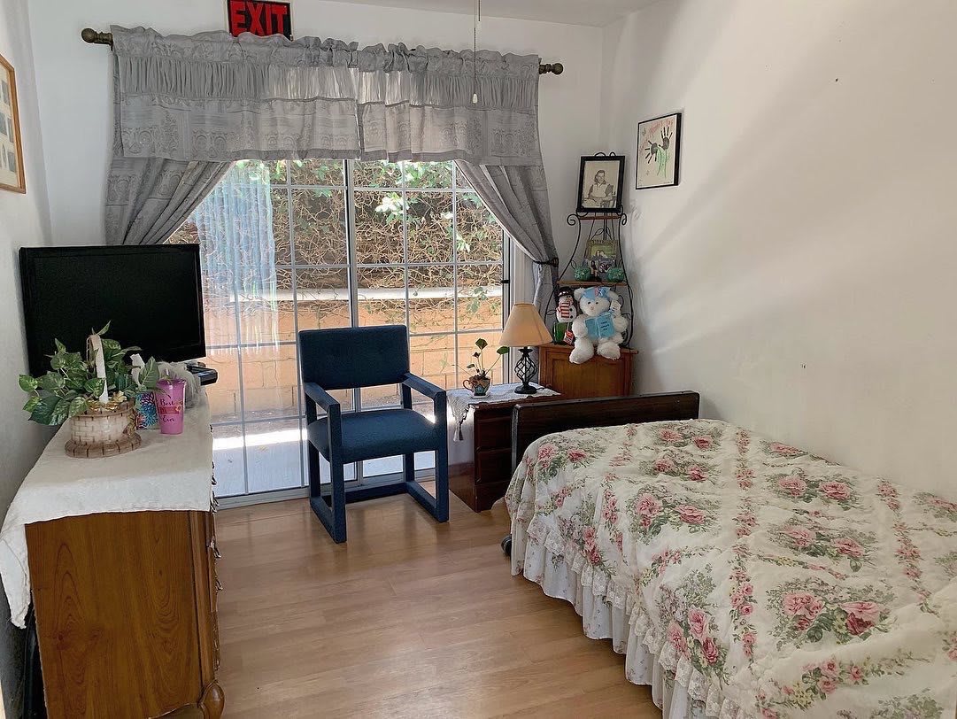 A cozy bedroom with a floral bedspread, a wooden chair, a lamp on a nightstand, and large windows covered by curtains.