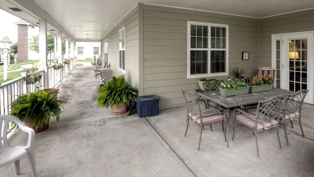 A covered outdoor patio area with a table, chairs, and potted plants along a concrete walkway.