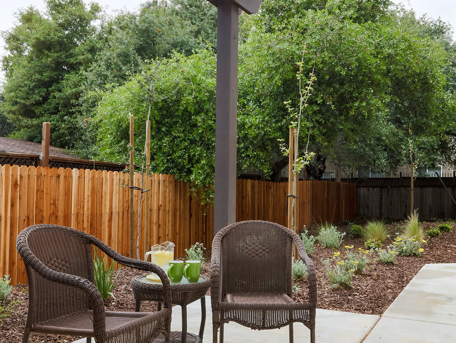 A cozy patio area with wicker chairs and a small table, surrounded by a garden and wooden fence.