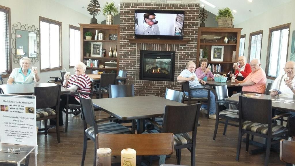 A cozy lounge area with people sitting around tables, a brick fireplace, and a television mounted above.