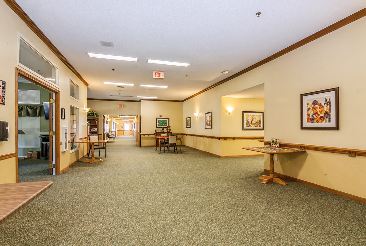 A spacious hallway with carpeted flooring, adorned with artwork and furnished with tables and chairs.
