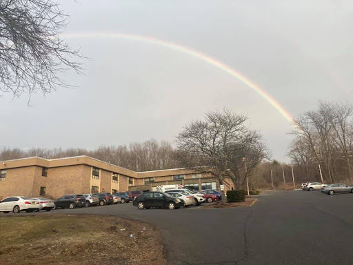 A beautiful rainbow arcs over a parking lot beside a building on a cloudy day.
