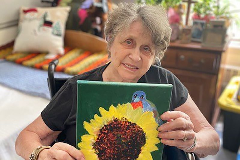 An elderly woman smiling while holding a painting of a sunflower with a bird perched on it.