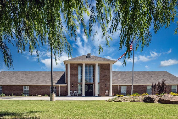 A brick building with a flag in front, surrounded by a green lawn and trees, under a blue sky.