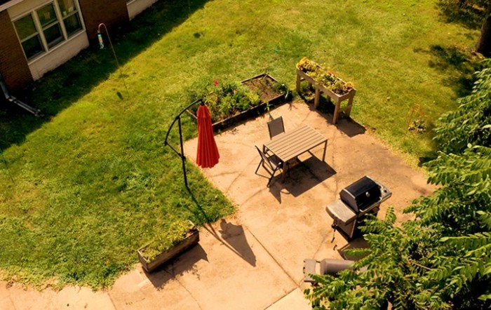Aerial view of a patio with a table, chairs, a red umbrella, grill, and planters in a backyard setting.