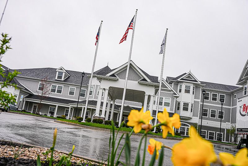 A large building with an entrance featuring three flagpoles and blooming yellow flowers in the foreground on a cloudy day.
