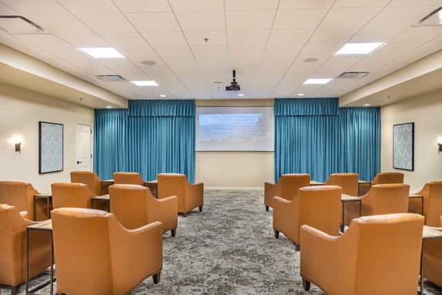 A small conference room with comfortable chairs facing a projector screen and surrounded by blue curtains.