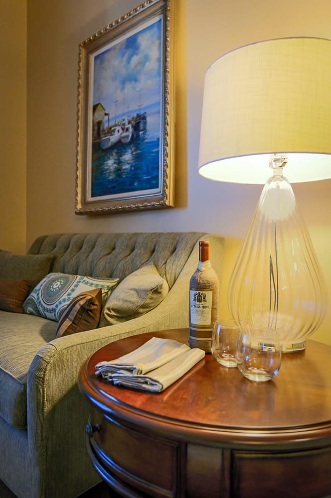 A cozy living room setup featuring a sofa, table with a bottle and glasses, and a warm lamp.