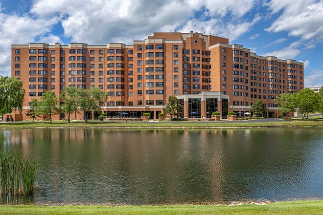 A large residential building with a lake in the foreground reflecting the structure and sky.