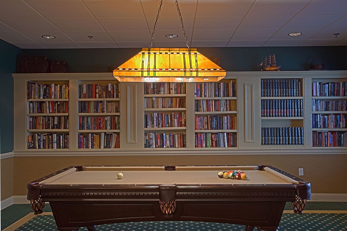 A billiards room with a pool table under a warm light fixture, surrounded by bookshelves filled with books.