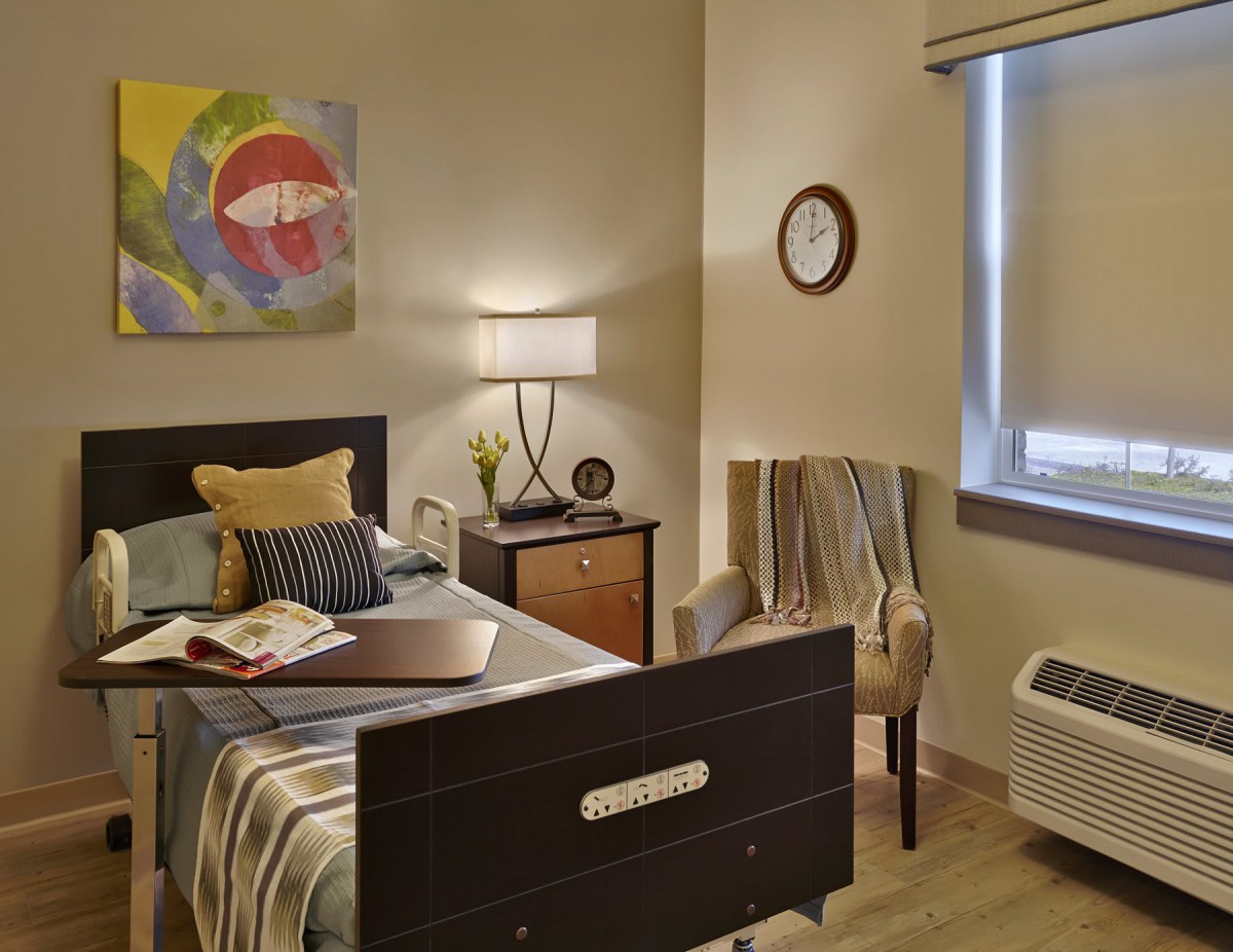 A cozy bedroom with modern decor, featuring a bed, side table with a lamp, colorful wall art, and an upholstered chair by the window.