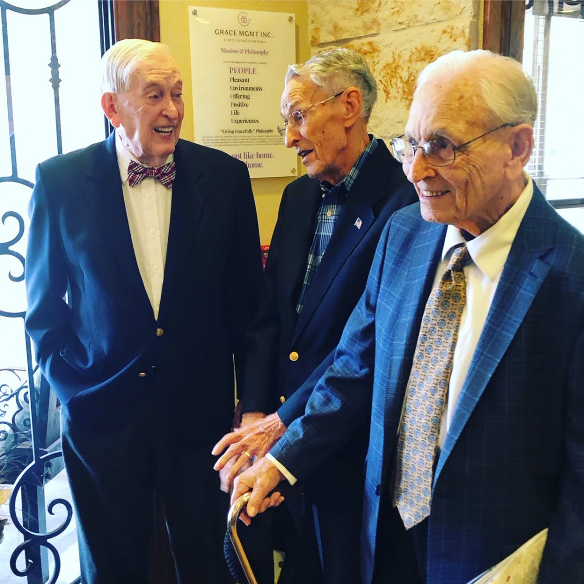 Three elderly men in suits engage in conversation indoors.