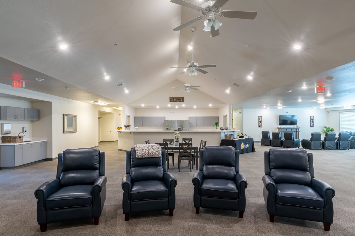 A spacious modern living room with four black recliners, a dining area in the background, and ceiling fans overhead.