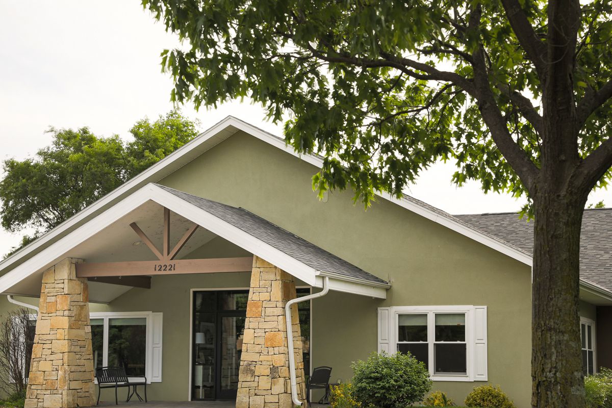 A quaint green house with a sloped roof and stone pillar under a tree.