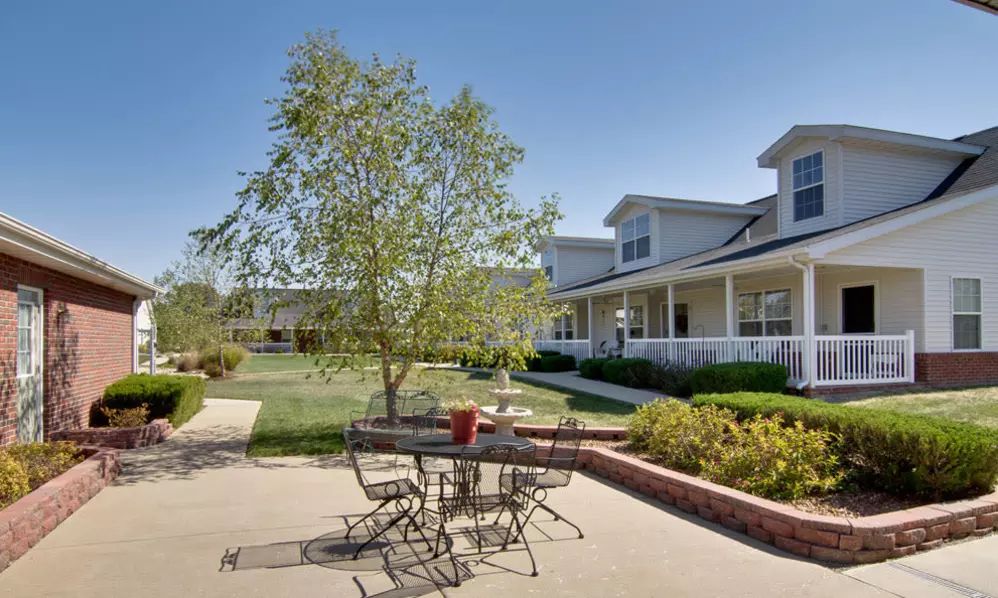 A sunny outdoor patio with a table and chairs, next to a garden and house.