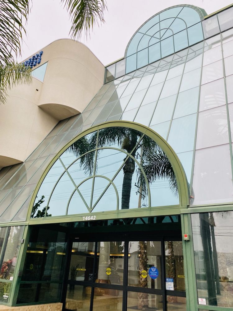 A modern shopping mall entrance with a glass facade and palm trees reflected in the windows.