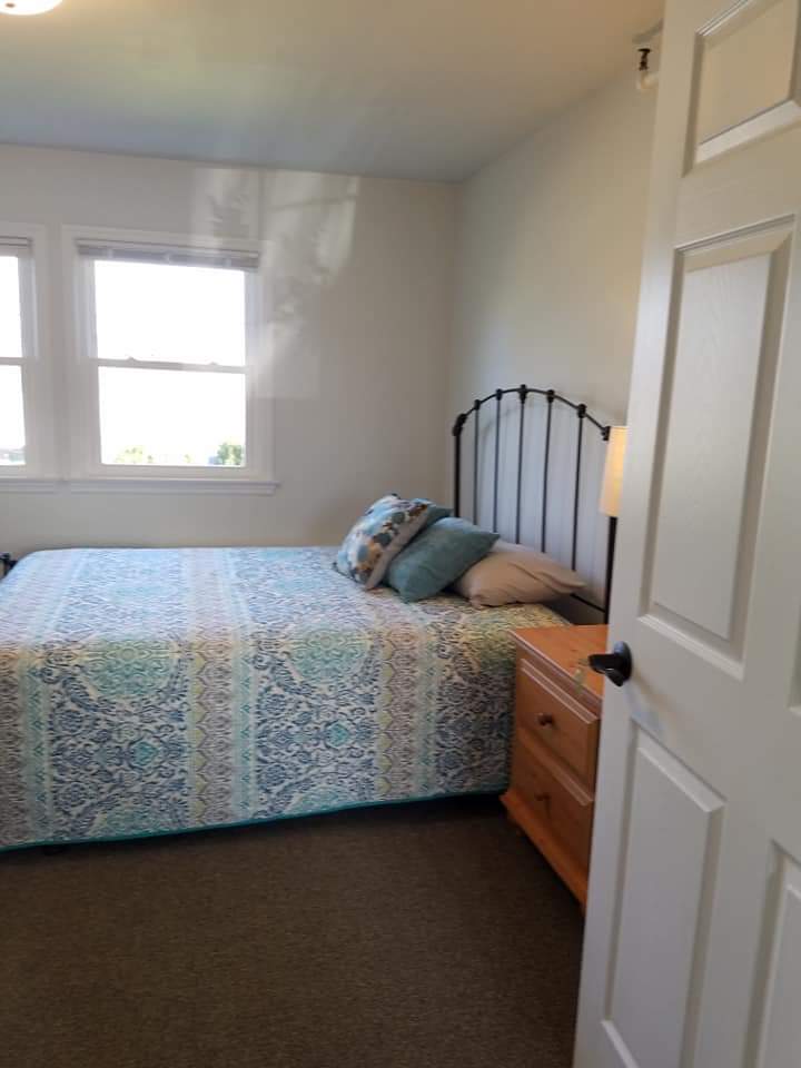 A cozy bedroom with a bed, nightstand, and windows letting in natural light.
